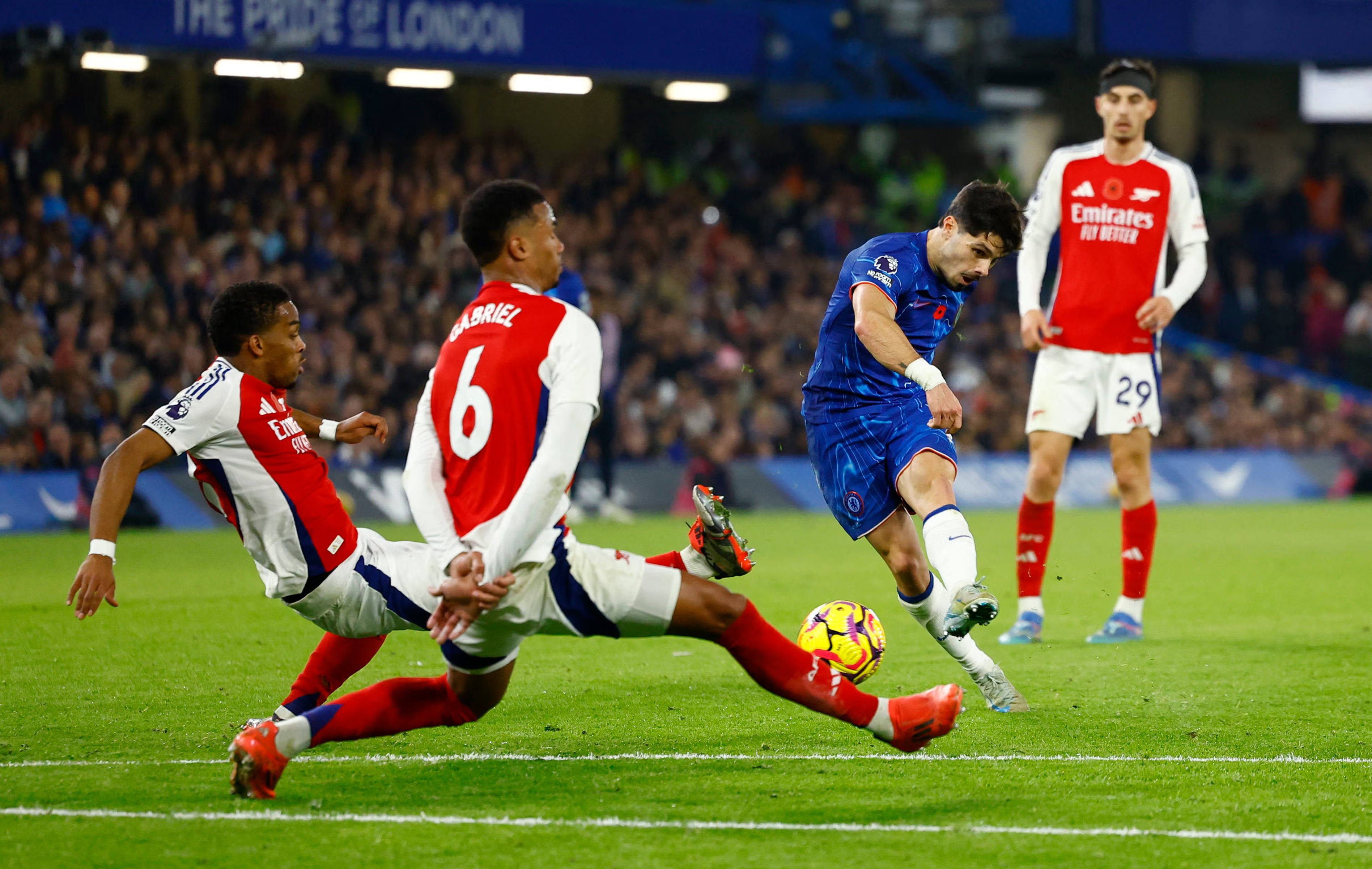 Chelsea player striking the ball while two Arsenal defenders attempt a block during a Premier League match at Stamford Bridge.